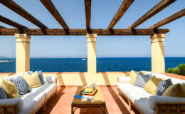 Cozy outdoor seating area with decorative pillows overlooking a serene blue sea, featuring a wooden pergola and a table with refreshments. Ideal for relaxation and enjoying coastal views.