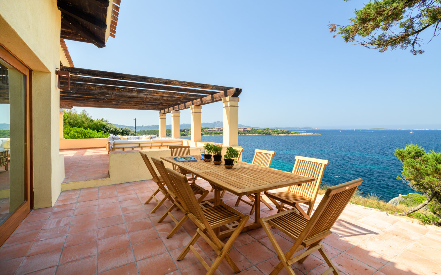 Outdoor dining area with wooden table and chairs overlooking a serene ocean view, featuring a sunny terrace and lush greenery. Ideal for relaxation and entertaining in a coastal setting. | Only on SopranoVillas