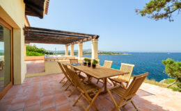 Outdoor dining area with wooden table and chairs overlooking a serene ocean view, featuring a sunny terrace and lush greenery. Ideal for relaxation and entertaining in a coastal setting.
