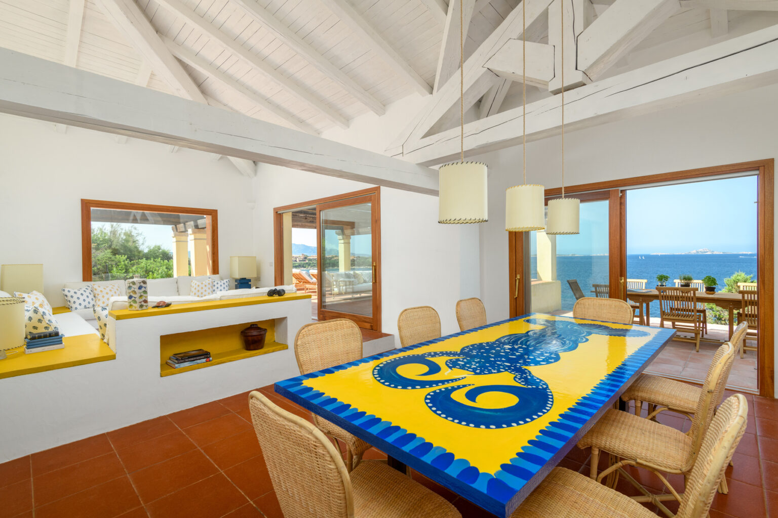 Bright and airy dining area featuring a vibrant yellow table with an octopus design, surrounded by woven chairs. The room boasts high white ceilings and large windows offering stunning ocean views. A cozy seating nook with decorative pillows and a modern aesthetic complements the inviting atmosphere. | Only on SopranoVillas