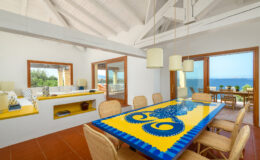 Bright and airy dining area featuring a vibrant yellow table with an octopus design, surrounded by woven chairs. The room boasts high white ceilings and large windows offering stunning ocean views. A cozy seating nook with decorative pillows and a modern aesthetic complements the inviting atmosphere.