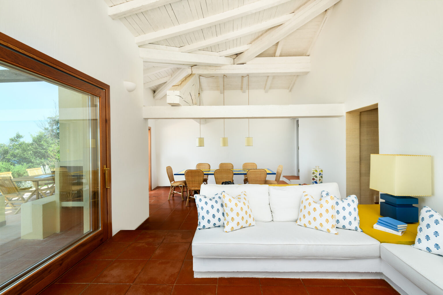 Modern living room with white sofa and colorful cushions, featuring high ceilings and natural light. The open layout connects to a dining area with wooden furniture, creating a spacious and inviting atmosphere. | Only on SopranoVillas