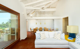 Modern living room with white sofa and colorful cushions, featuring high ceilings and natural light. The open layout connects to a dining area with wooden furniture, creating a spacious and inviting atmosphere.