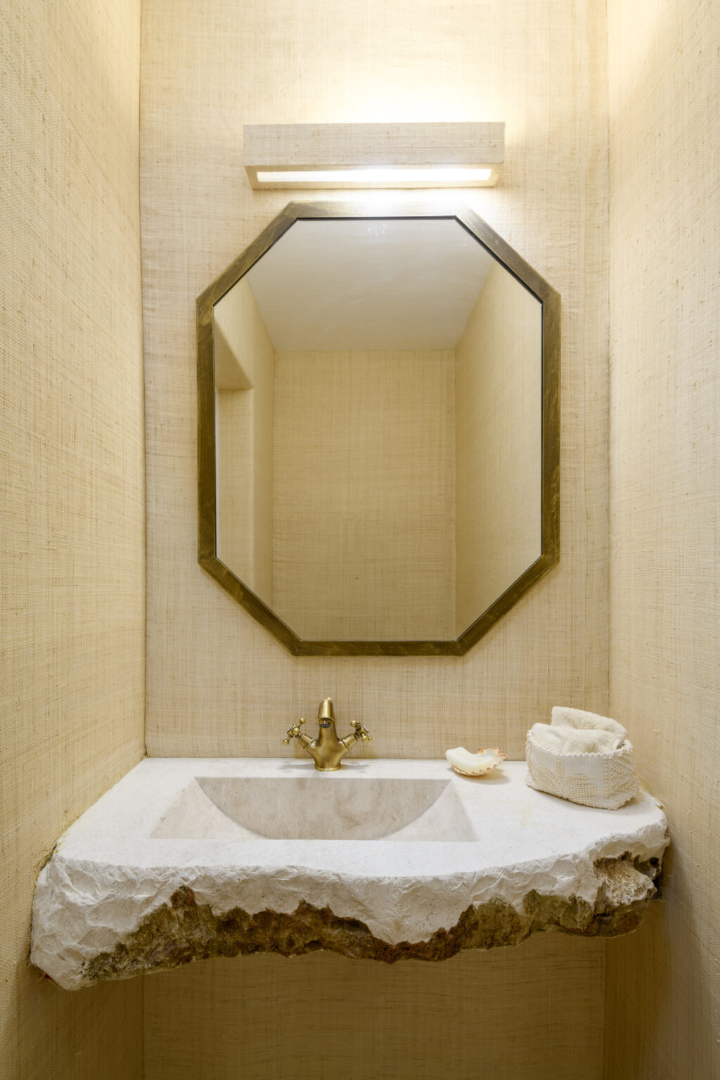 Modern bathroom design featuring a unique stone sink with a brass faucet, an octagonal mirror, and textured beige walls. The space is elegantly illuminated and decorated with a decorative shell and a soft towel holder. | Only on SopranoVillas