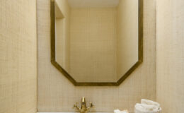 Modern bathroom design featuring a unique stone sink with a brass faucet, an octagonal mirror, and textured beige walls. The space is elegantly illuminated and decorated with a decorative shell and a soft towel holder.