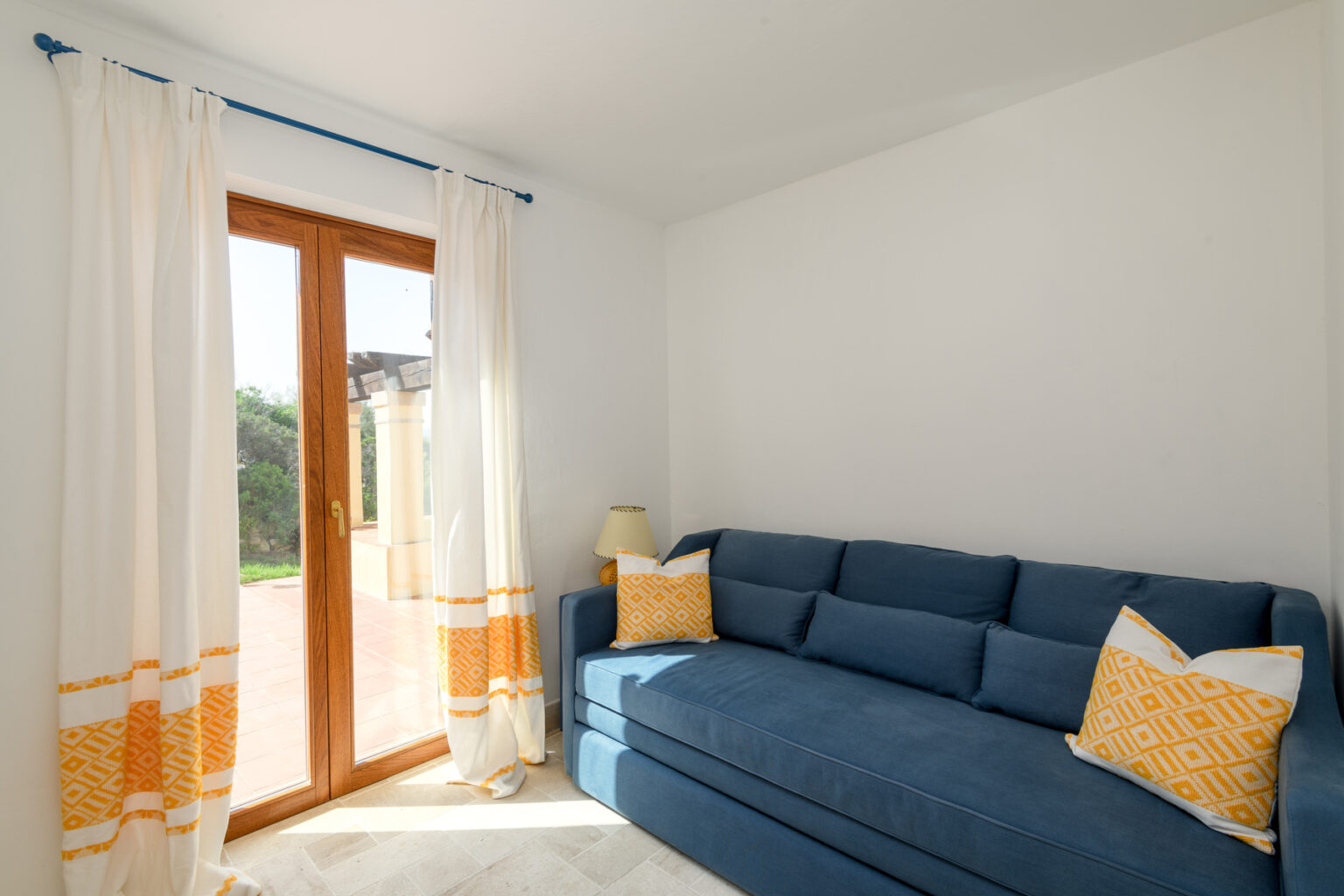 Cozy living room featuring a blue sofa with yellow accent pillows, white walls, and large windows with sheer curtains, allowing natural light to brighten the space. | Only on SopranoVillas