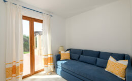 Cozy living room featuring a blue sofa with yellow accent pillows, white walls, and large windows with sheer curtains, allowing natural light to brighten the space.