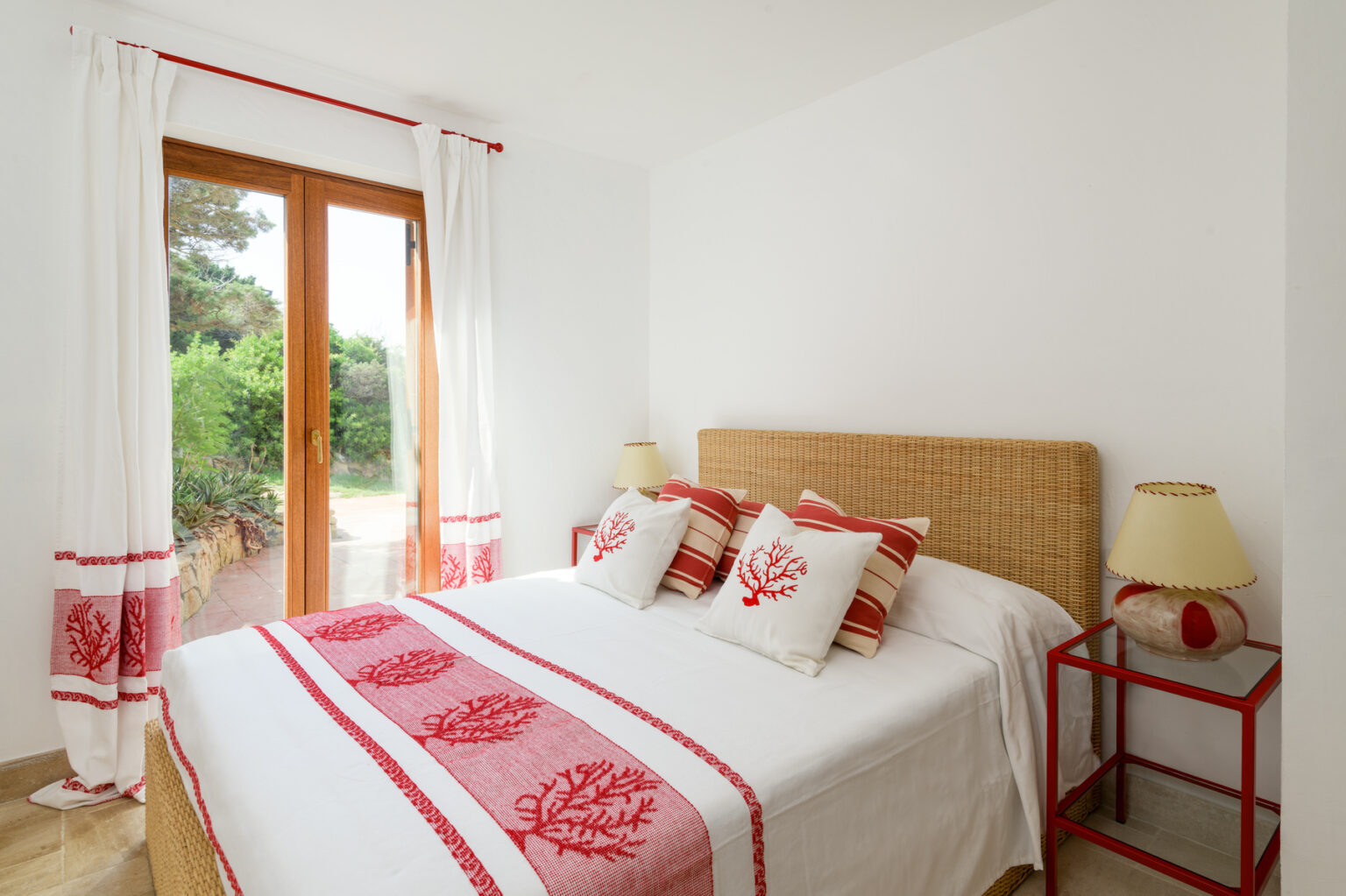 Cozy bedroom featuring a double bed with decorative red and white pillows, natural wicker headboard, and matching bedside lamps. Large windows offer a view of a green garden, enhancing the bright and airy atmosphere. | Only on SopranoVillas