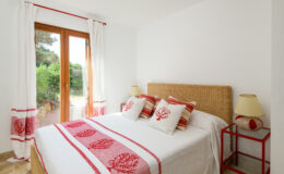 Cozy bedroom featuring a double bed with decorative red and white pillows, natural wicker headboard, and matching bedside lamps. Large windows offer a view of a green garden, enhancing the bright and airy atmosphere.