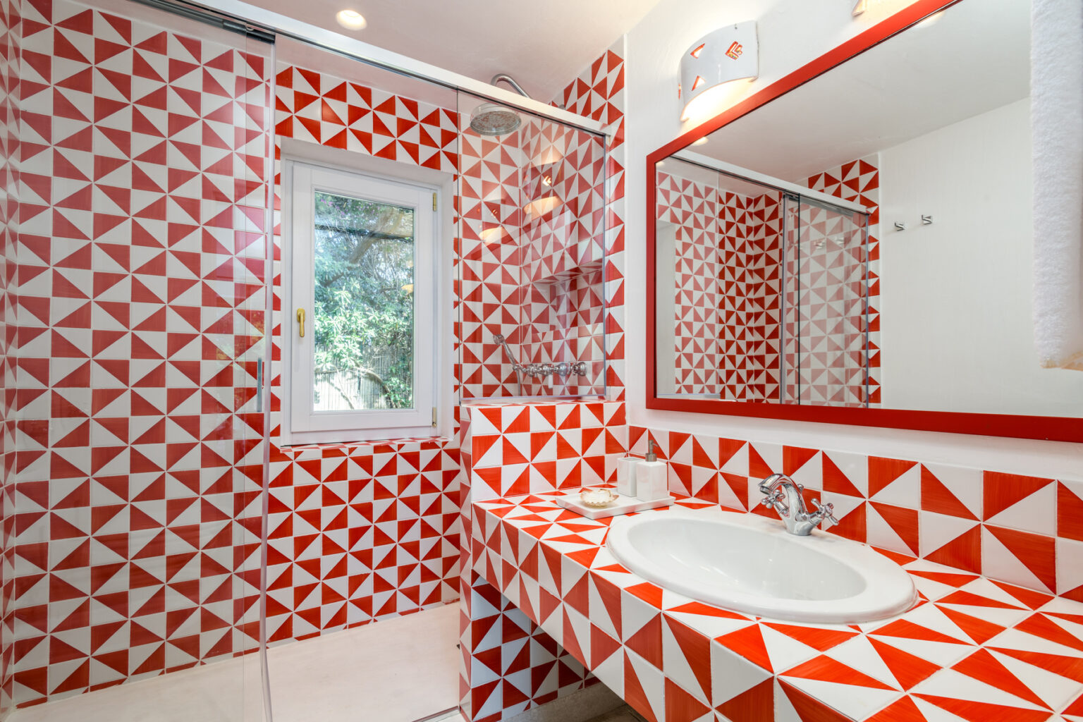 Bright and modern bathroom featuring bold red and white geometric patterned tiles on walls and countertop, a sleek white sink, and a large mirror reflecting natural light from a window. | Only on SopranoVillas