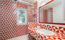 Bright and modern bathroom featuring bold red and white geometric patterned tiles on walls and countertop, a sleek white sink, and a large mirror reflecting natural light from a window.