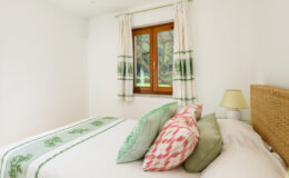 Bright and airy bedroom featuring a cozy bed with decorative pillows in green and pink hues, a wooden window with natural light, and light-colored curtains. Ideal for showcasing modern interior design and comfort.