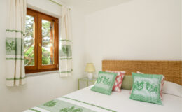 Cozy bedroom featuring a comfortable bed with green and pink decorative pillows, natural light streaming through a wooden window, and stylish curtains, creating a serene and inviting atmosphere.