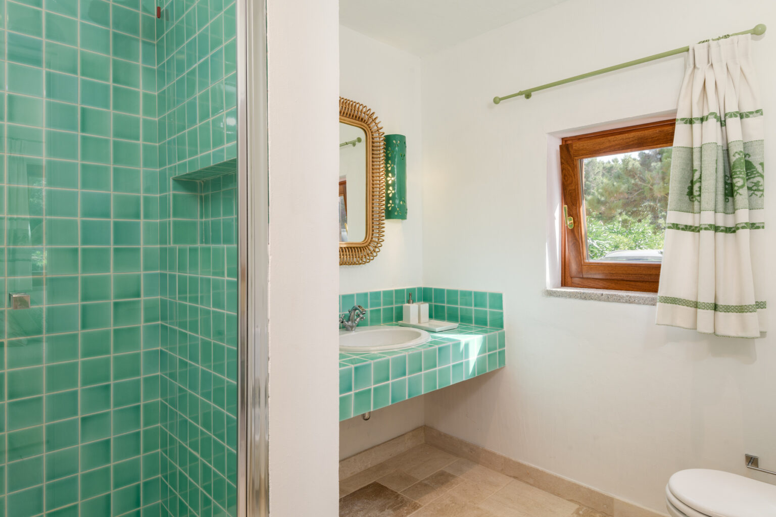 Bright and modern bathroom featuring turquoise tiled walls, a glass shower, and a matching sink area, complemented by a wooden window with white and green curtains. | Only on SopranoVillas