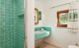 Bright and modern bathroom featuring turquoise tiled walls, a glass shower, and a matching sink area, complemented by a wooden window with white and green curtains.