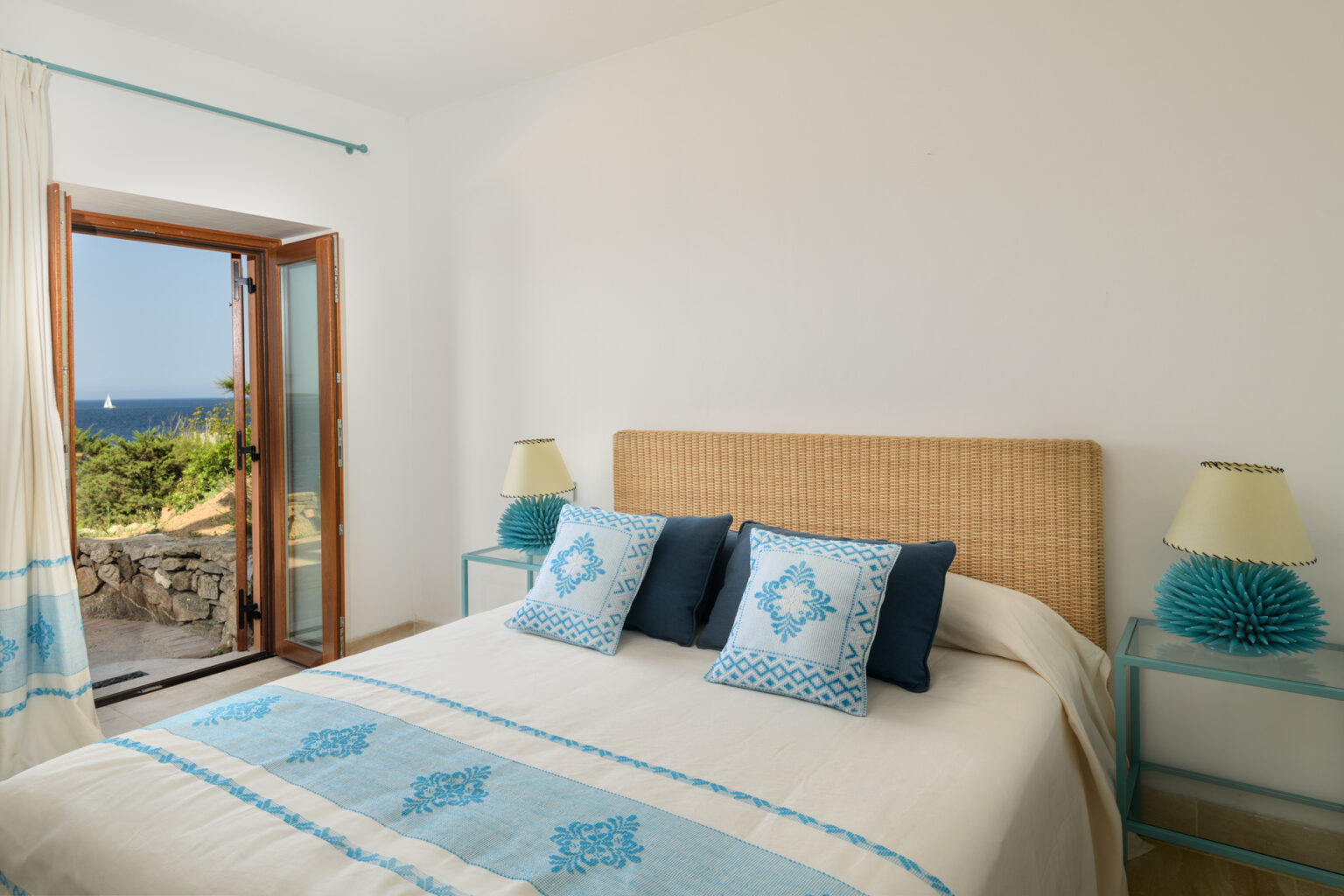 Cozy bedroom with a queen-sized bed adorned with blue and white decorative pillows, natural woven headboard, and bedside lamps. Large window offers a scenic view of the outdoors, enhancing the bright and airy atmosphere. Ideal for relaxation and comfort. | Only on SopranoVillas