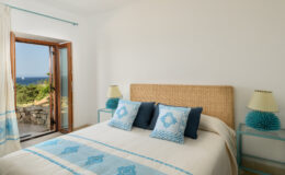 Cozy bedroom with a queen-sized bed adorned with blue and white decorative pillows, natural woven headboard, and bedside lamps. Large window offers a scenic view of the outdoors, enhancing the bright and airy atmosphere. Ideal for relaxation and comfort.