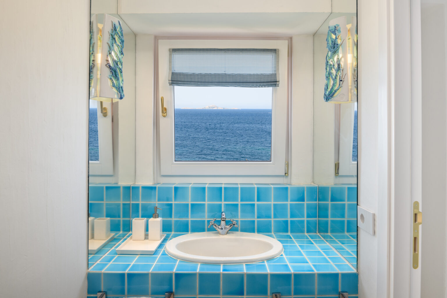 Bright and airy bathroom featuring blue tiled walls and a circular bathtub, with large windows offering a stunning ocean view. | Only on SopranoVillas