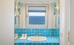Bright and airy bathroom featuring blue tiled walls and a circular bathtub, with large windows offering a stunning ocean view.