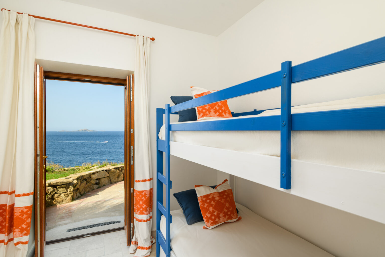 Cozy bedroom with blue bunk beds and bright orange decorative pillows, featuring a large window that opens to a stunning ocean view. | Only on SopranoVillas