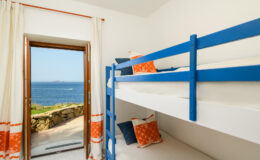 Cozy bedroom with blue bunk beds and bright orange decorative pillows, featuring a large window that opens to a stunning ocean view.
