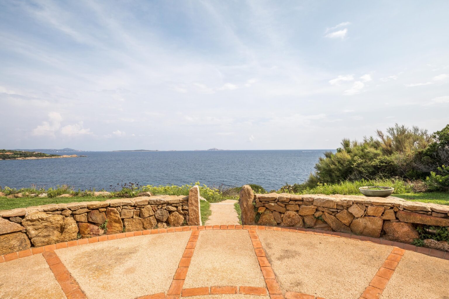 Scenic coastal view from a stone pathway overlooking the ocean, with lush greenery and distant islands under a partly cloudy sky. | Only on SopranoVillas