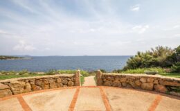 Scenic coastal view from a stone pathway overlooking the ocean, with lush greenery and distant islands under a partly cloudy sky.
