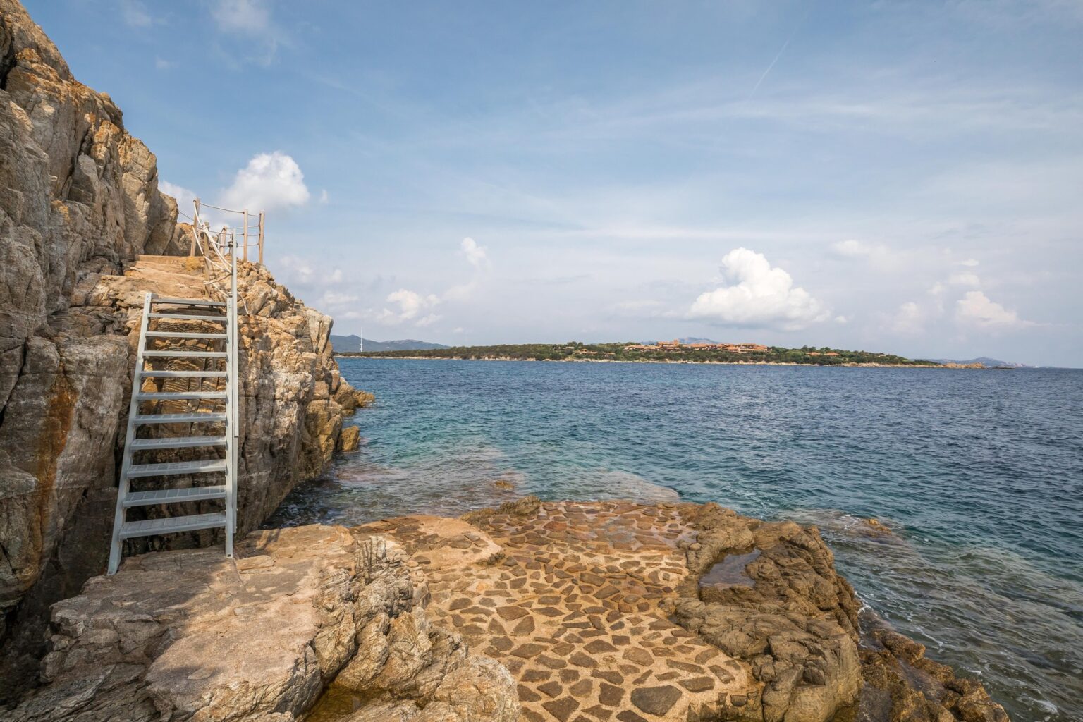 Scenic view of rocky coastline with a metal staircase leading down to the calm blue sea, under a partly cloudy sky, featuring a distant shoreline with greenery and buildings. | Only on SopranoVillas