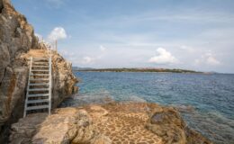 Scenic view of rocky coastline with a metal staircase leading down to the calm blue sea, under a partly cloudy sky, featuring a distant shoreline with greenery and buildings.