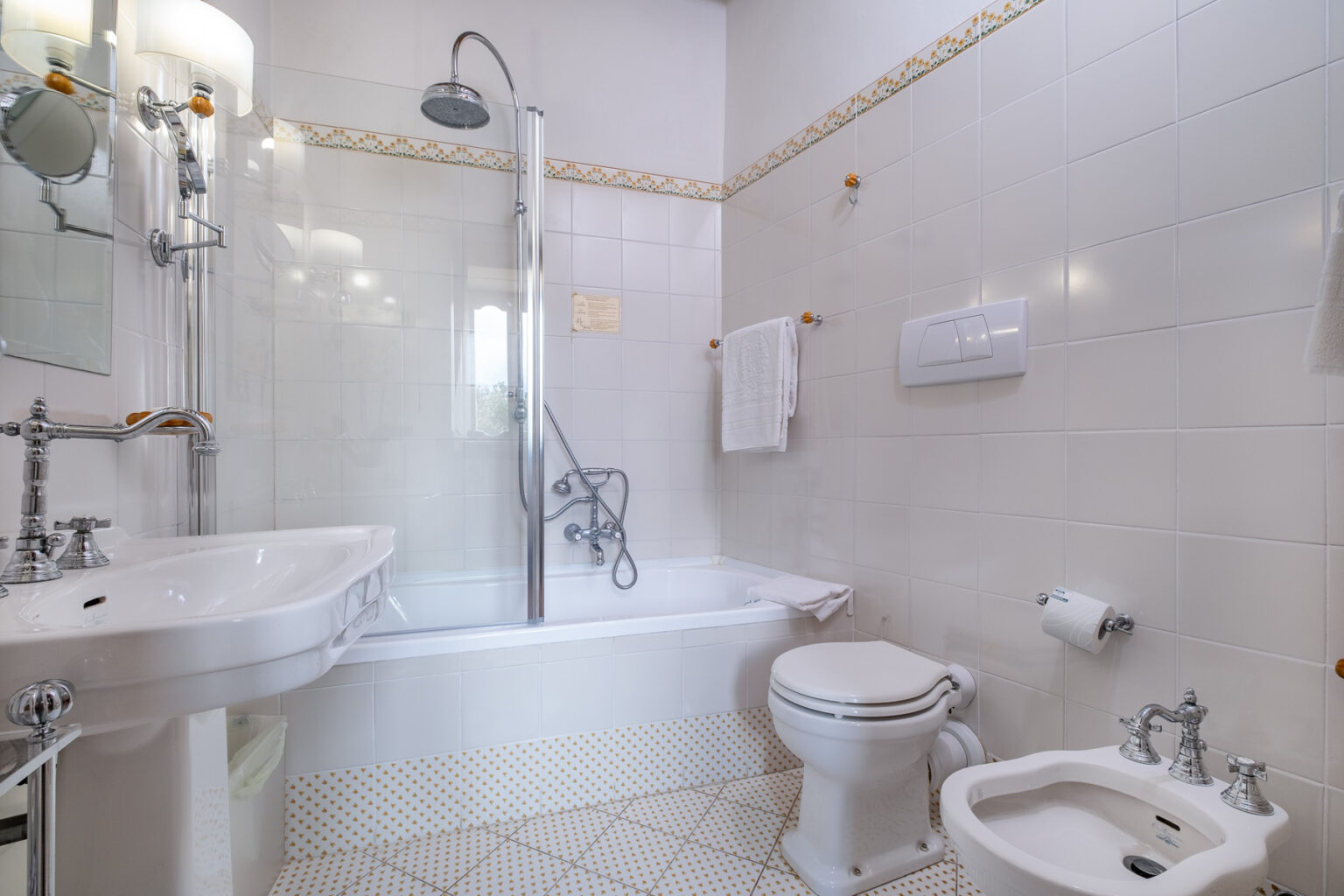 Modern bathroom featuring a bathtub with a shower, white tiled walls, a pedestal sink, a toilet, and a bidet. The space is well-lit with decorative accents and neatly arranged towels. Ideal for showcasing contemporary bathroom design. | Only on SopranoVillas