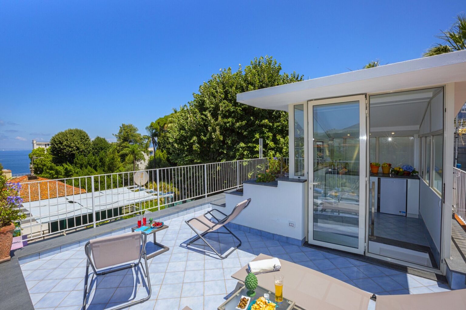 Sunny terrace with lounge chairs overlooking the ocean, featuring a glass door leading to a modern interior space surrounded by lush greenery. Ideal for relaxation and outdoor living. | Only on SopranoVillas