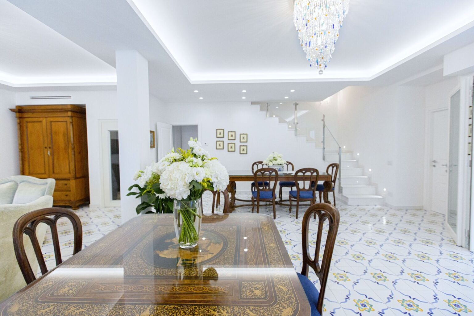 Modern dining room featuring a wooden dining table with elegant chairs, a vase of flowers, and a stylish chandelier. The space is bright and airy, with white walls and decorative tiles, creating a welcoming atmosphere. | Only on SopranoVillas