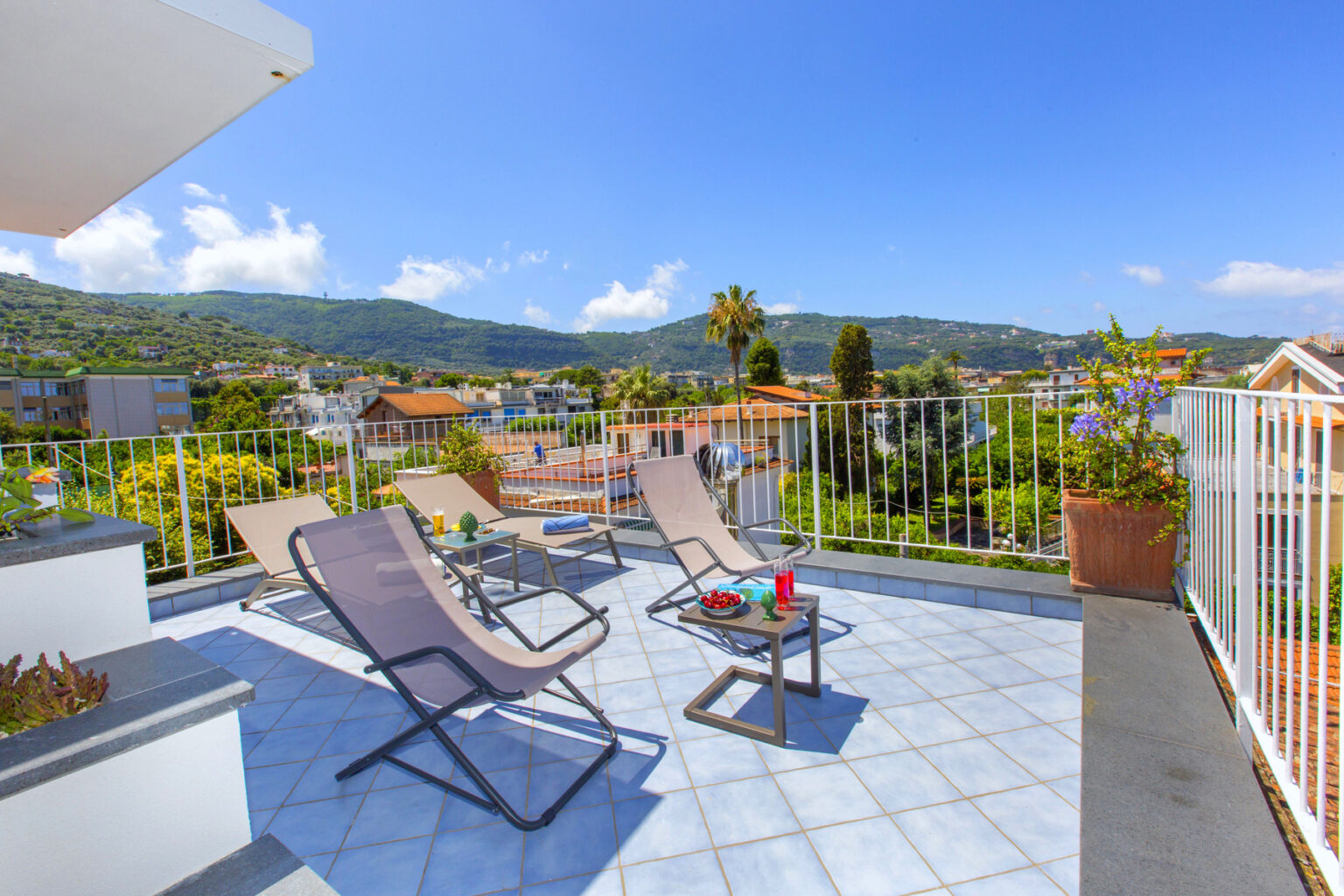 Spacious balcony with lounge chairs and a table, offering a panoramic view of a scenic landscape with mountains and houses under a clear blue sky. Ideal for relaxation and outdoor dining. | Only on SopranoVillas