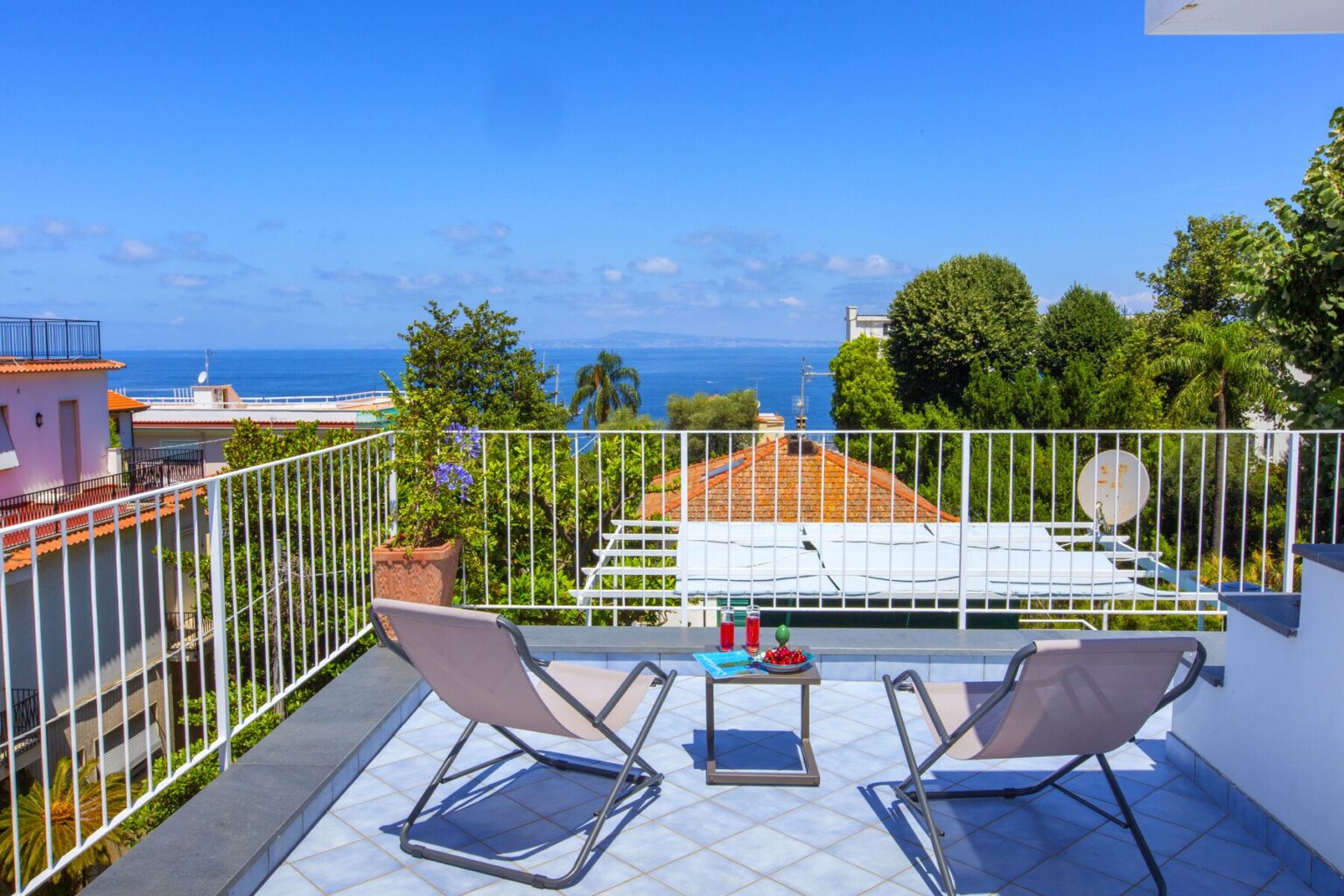 A sunny terrace with two lounge chairs overlooking a scenic ocean view, surrounded by greenery and colorful rooftops, ideal for relaxation and leisure. | Only on SopranoVillas