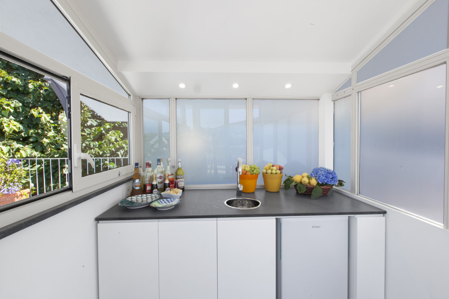 Bright and modern kitchen space featuring a sleek countertop, colorful fruit bowl, and various drinks on display. Large windows provide ample natural light, enhancing the airy atmosphere. Ideal for entertaining and cooking. | Only on SopranoVillas
