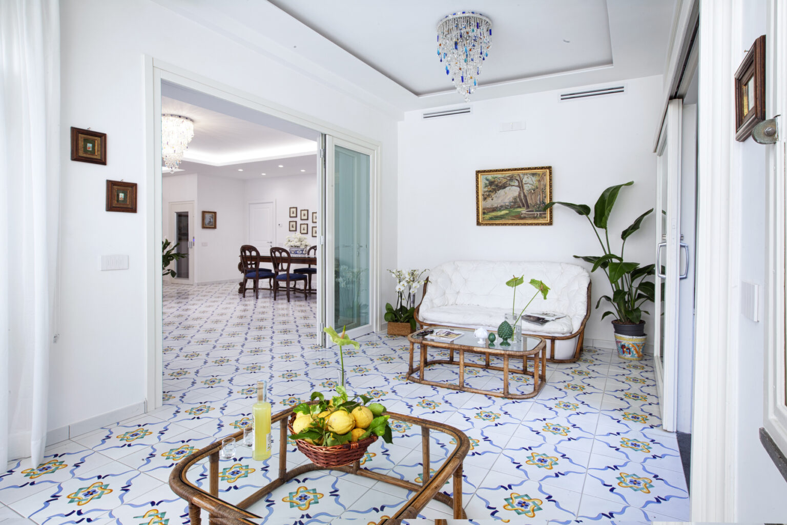 Bright and airy living room featuring decorative tiled flooring, a cozy white sofa, and a glass coffee table with a fruit basket. Elegant chandeliers and framed artwork enhance the stylish interior, leading to a dining area in the background. Lush greenery adds a touch of nature to the space. | Only on SopranoVillas