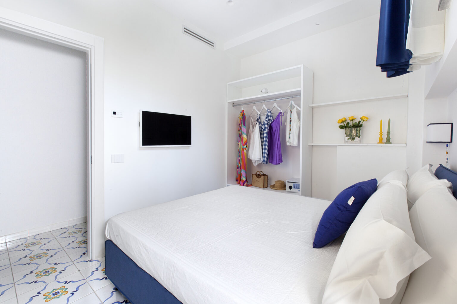 Bright and airy bedroom featuring a queen-sized bed with decorative pillows, a wall-mounted TV, and a stylish wardrobe displaying colorful clothing. The room has white walls and patterned tile flooring, creating a modern and inviting atmosphere. | Only on SopranoVillas