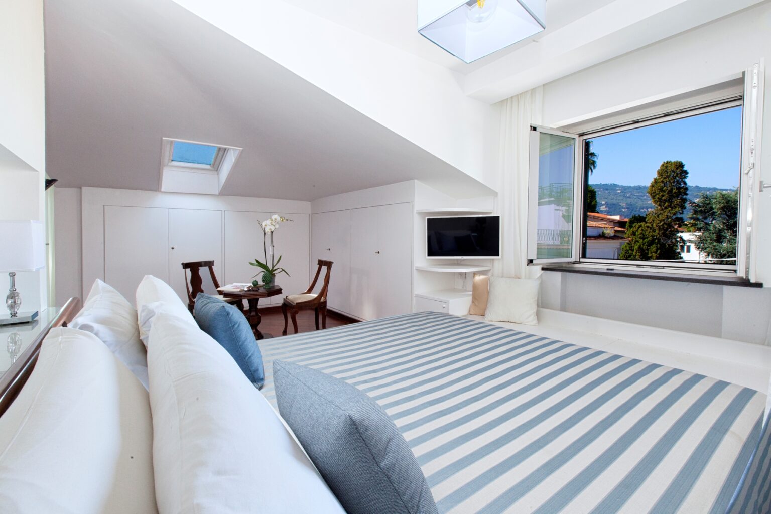 Modern bedroom interior featuring a striped bedspread, large windows with scenic views, and a cozy seating area with two chairs. Bright, minimalist design with natural light streaming in. | Only on SopranoVillas
