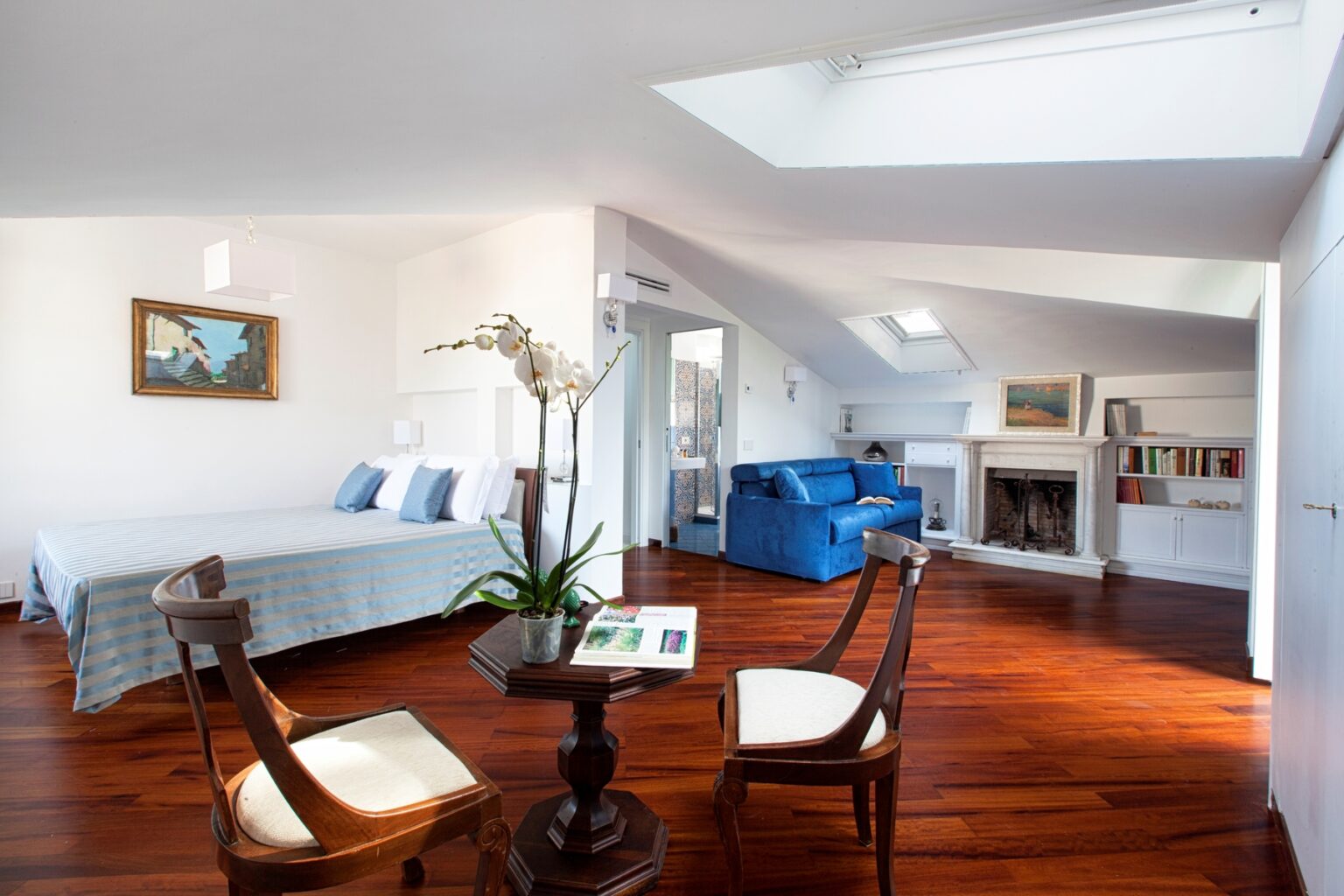 Spacious and elegantly designed attic bedroom featuring a comfortable bed with blue striped bedding, a cozy blue sofa, and hardwood flooring. The room includes a decorative fireplace, a small wooden table with a plant, and natural light from skylights. Art decorates the walls, enhancing the inviting atmosphere. | Only on SopranoVillas