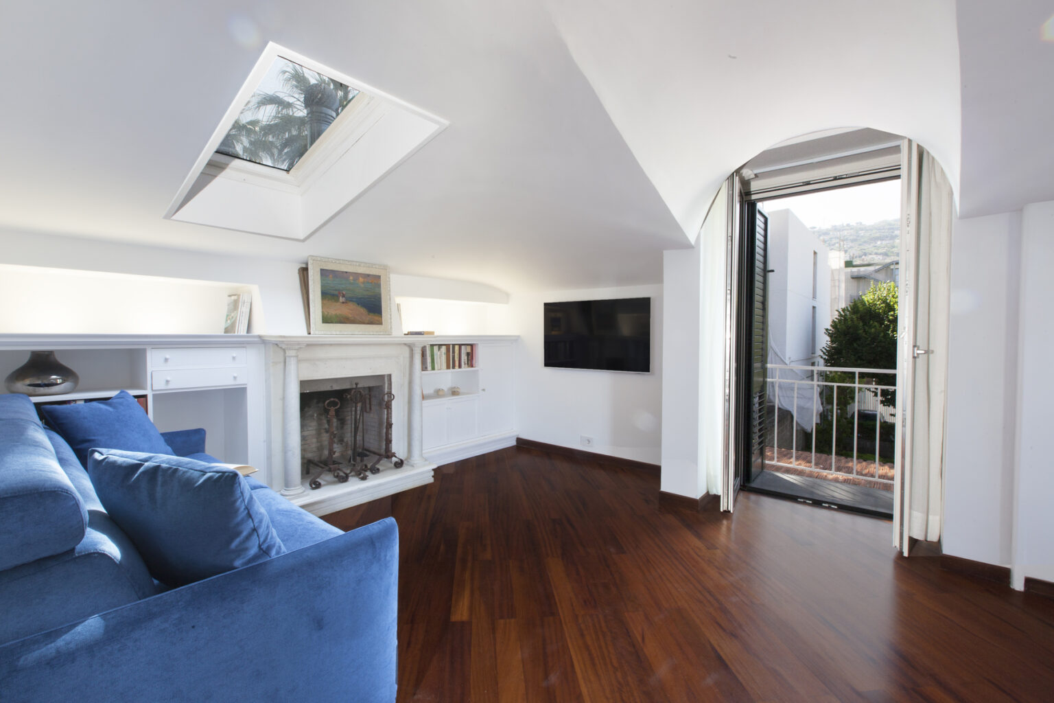 Spacious living room featuring a blue sofa, wooden flooring, and a fireplace, with a skylight and balcony door leading to an outdoor area. The room is well-lit with natural light and includes shelves with books and artwork. | Only on SopranoVillas
