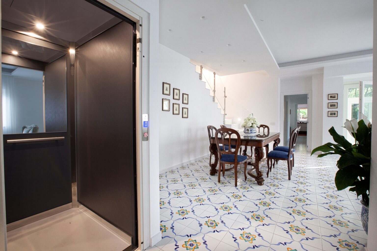 Modern interior featuring a residential elevator entrance leading into a stylish dining area with a wooden table and blue upholstered chairs, surrounded by decorative tiled flooring and elegant wall art. | Only on SopranoVillas