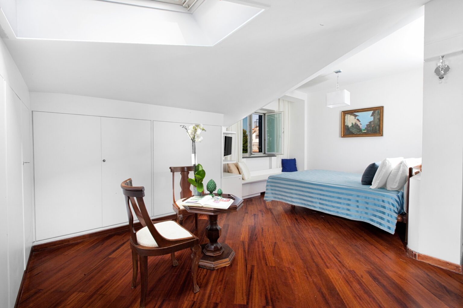Bright and airy bedroom featuring a blue bedspread, hardwood flooring, and a wooden chair. The room includes a small table with decorative items and a window providing natural light, creating a cozy and inviting atmosphere. | Only on SopranoVillas