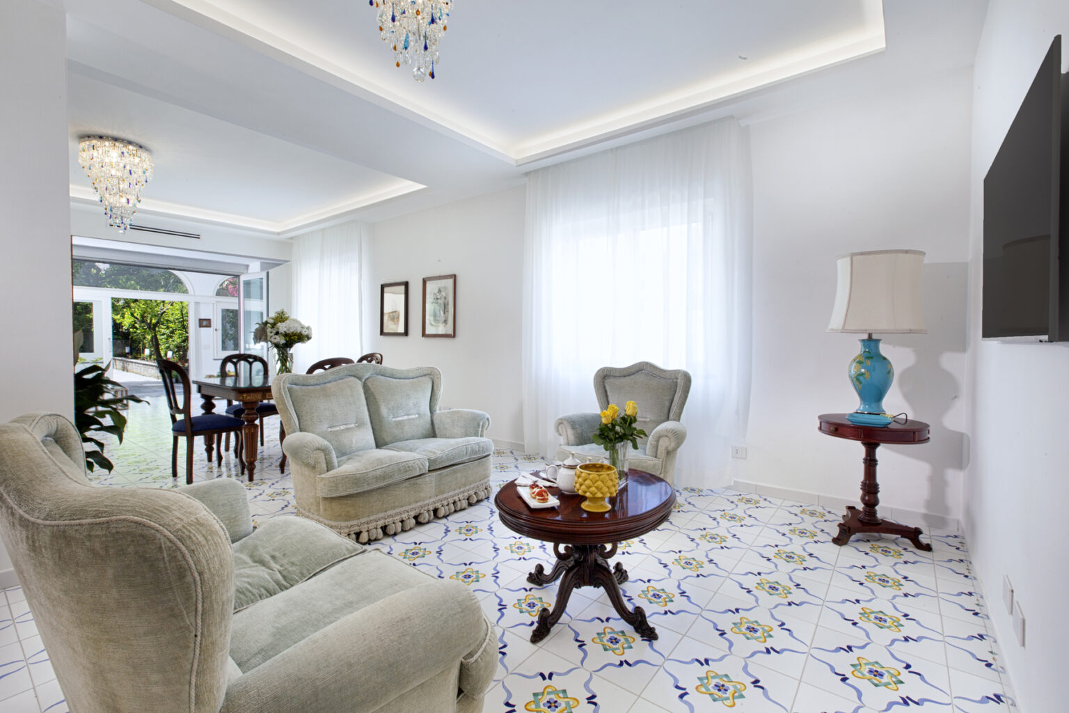 Bright and airy living room featuring elegant furniture, including a plush green sofa and armchairs, a decorative wooden coffee table, and a stylish lamp. The space is enhanced by large windows allowing natural light, floral decor, and a modern TV, set against a vibrant tiled floor. | Only on SopranoVillas