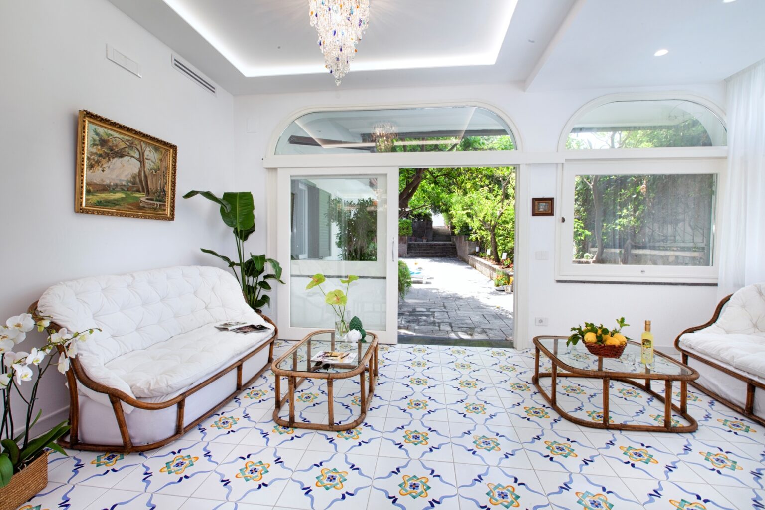 Bright and airy living room featuring white sofas, a glass coffee table, and a decorative chandelier, with large windows offering views of a lush outdoor garden and intricate tiled flooring. | Only on SopranoVillas