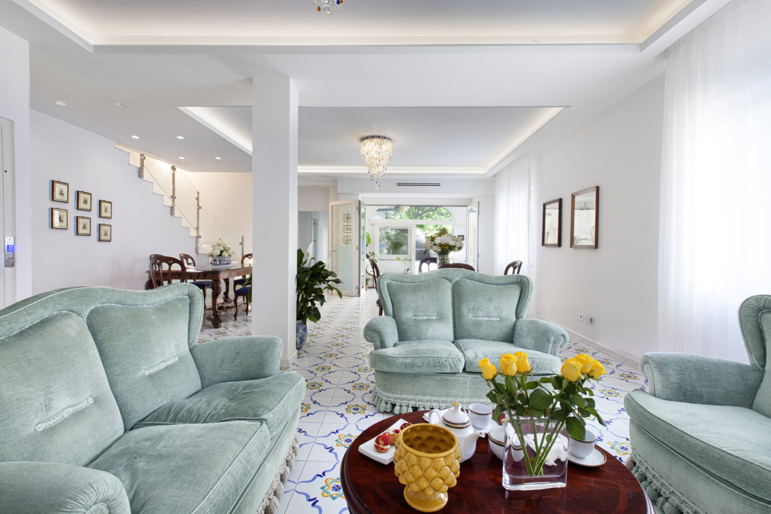 Spacious modern living room featuring light green sofas, a round wooden coffee table with a floral arrangement, and bright natural lighting from large windows. The open layout connects to a dining area in the background, enhancing the airy atmosphere. | Only on SopranoVillas