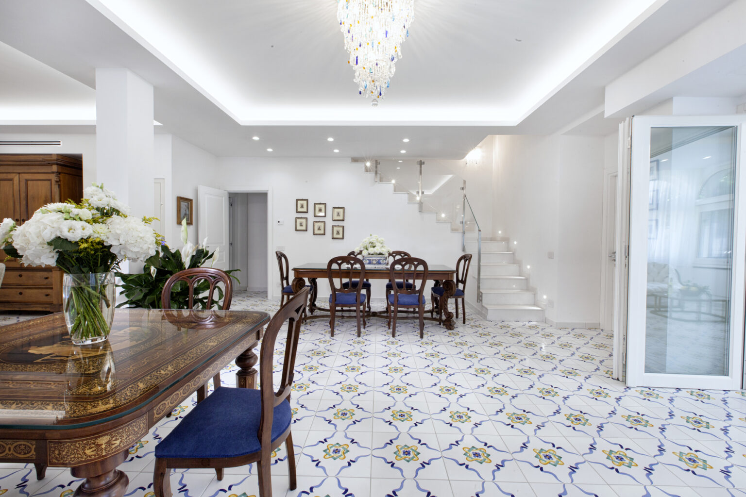 Bright and spacious modern dining room featuring a wooden dining table with blue upholstered chairs, elegant chandelier, and decorative tile flooring. The room includes a staircase and large windows for natural light, creating an inviting atmosphere. | Only on SopranoVillas