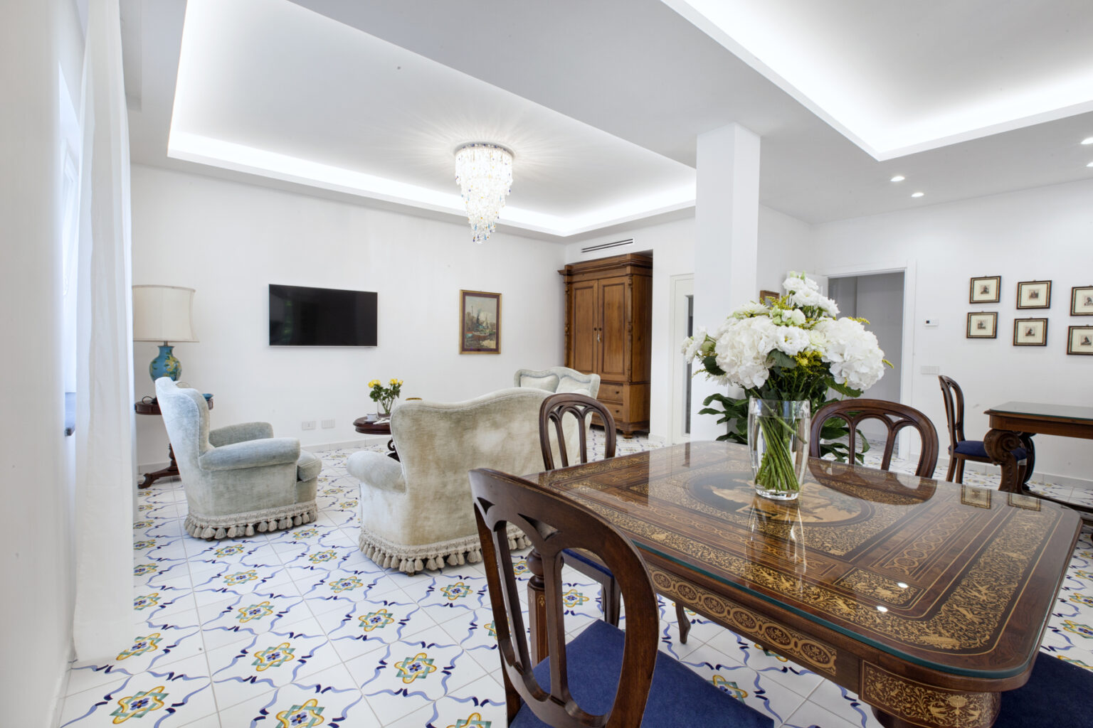 Bright and elegant living room featuring a wooden dining table, plush seating, decorative floral arrangements, and intricate tiled flooring, complemented by modern lighting and wall art. | Only on SopranoVillas