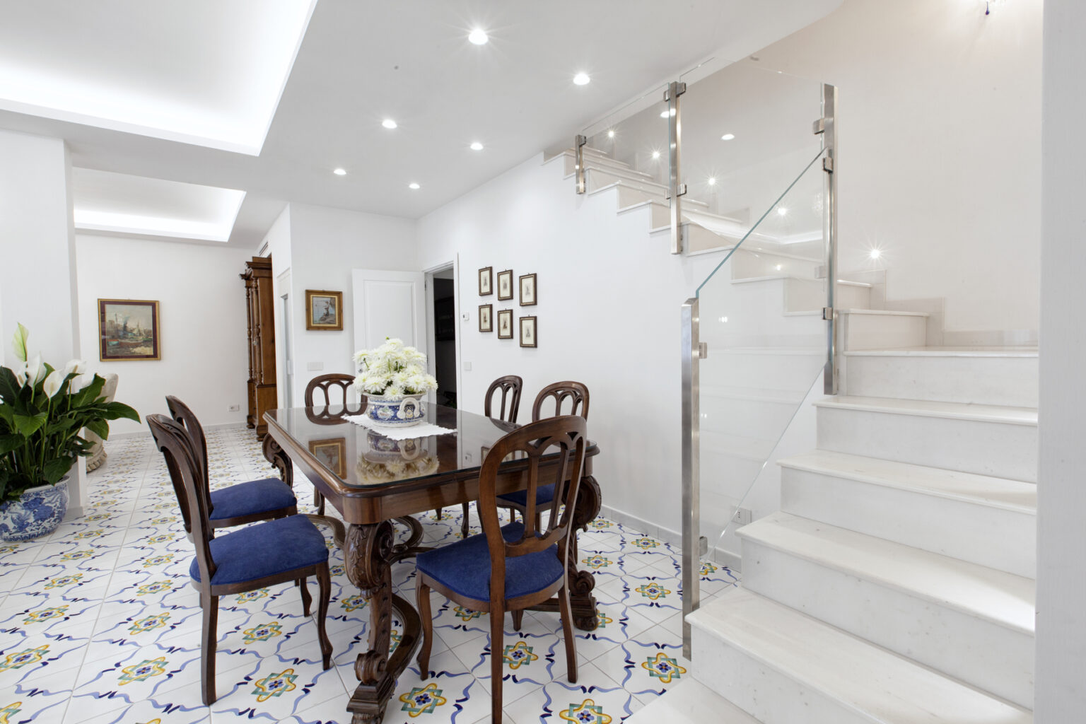 Bright and inviting interior of a modern dining area featuring a wooden table with blue upholstered chairs, decorative floral centerpiece, and elegant wall art. A staircase with glass railing leads to the upper level, complemented by stylish tile flooring. | Only on SopranoVillas