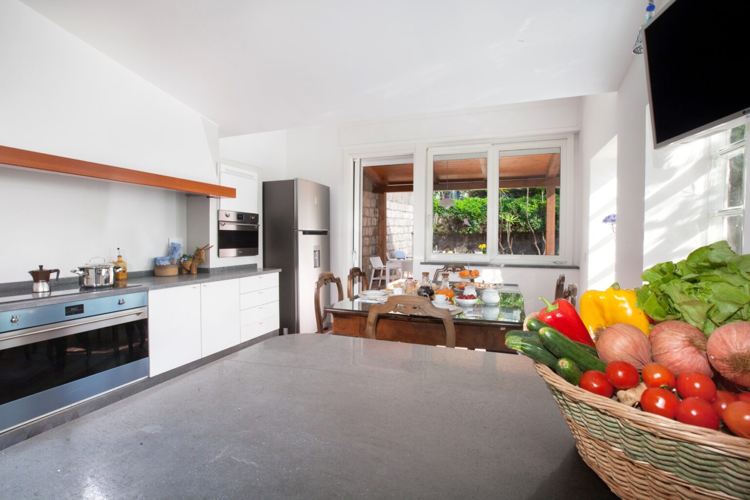 Modern kitchen with sleek white cabinetry, a large island countertop, and a view of a dining area. A basket of fresh vegetables and fruits adds color and vibrancy to the space, enhancing the inviting atmosphere. Natural light floods in through large windows, creating a warm and welcoming environment. | Only on SopranoVillas