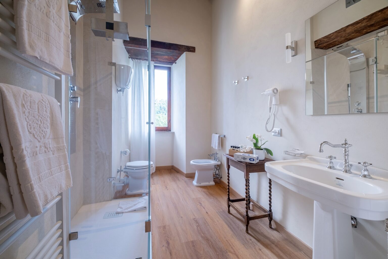 Modern bathroom featuring a glass shower, elegant vanity with vintage-style fixtures, a toilet, and natural light from a window. Soft towels are displayed, and a small table holds toiletries and a decorative plant, enhancing the tranquil atmosphere. | Only on SopranoVillas